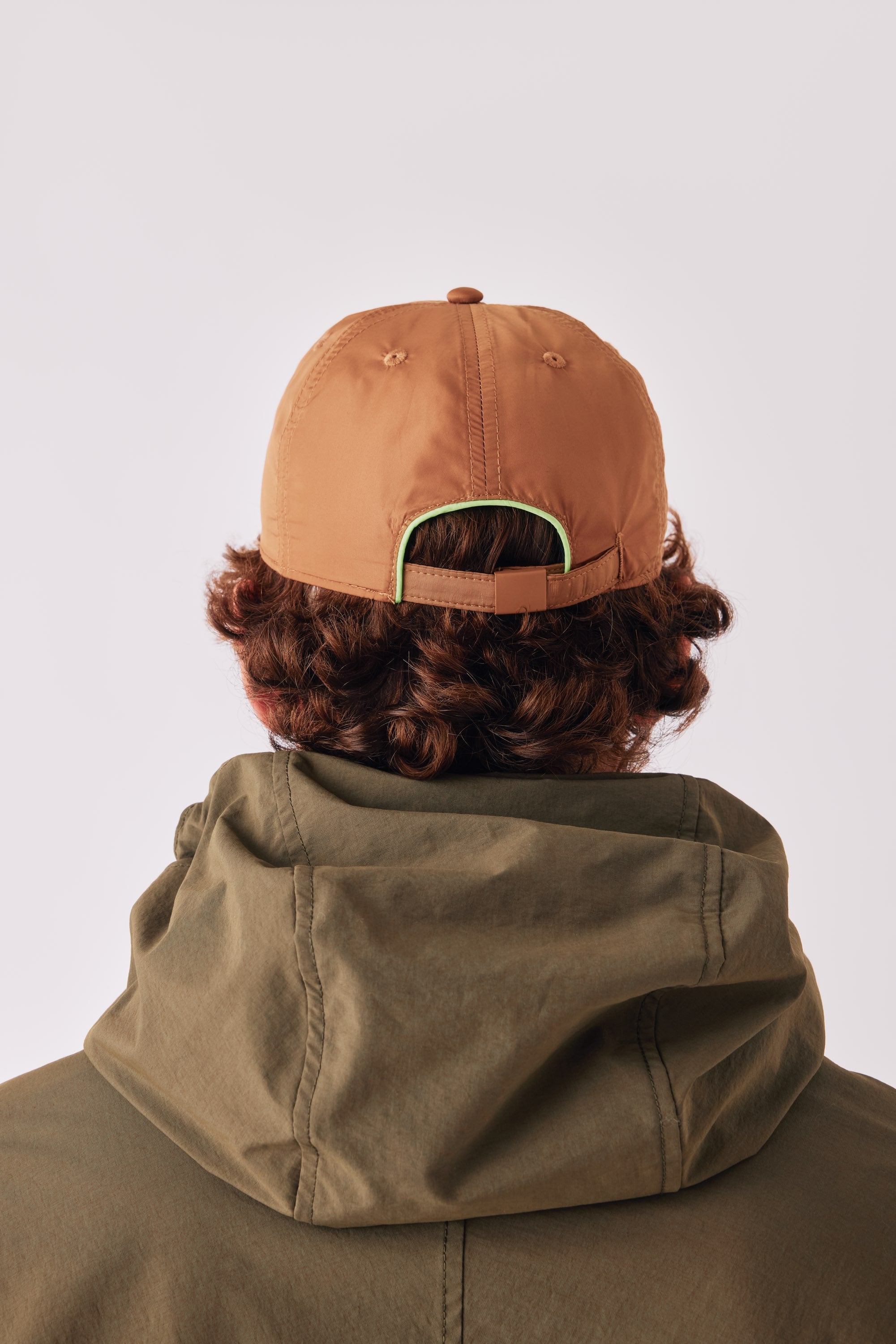 Adsum FW26 — back of a tan cap with a green trim strap, worn with an olive hood