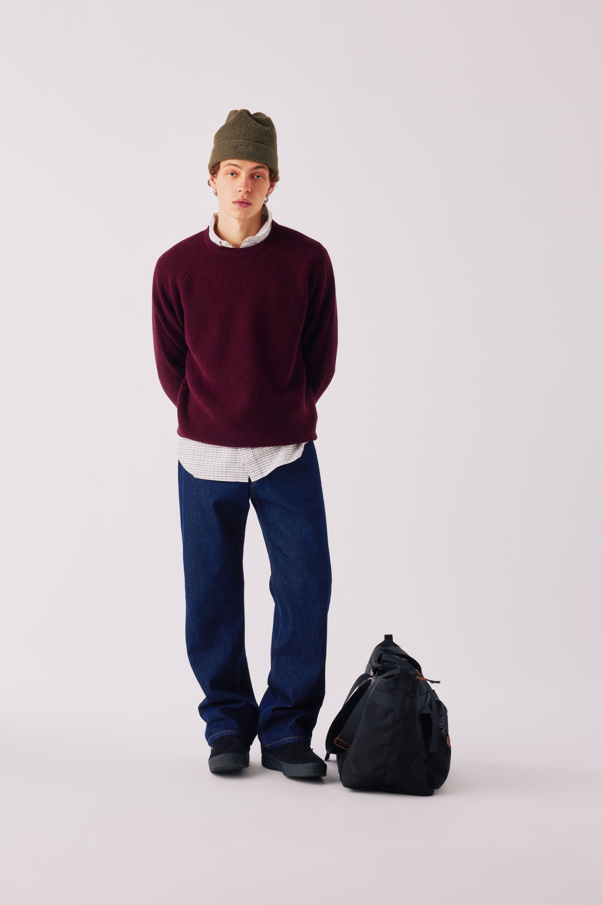 Adsum FW26 — model in a maroon sweater and dark denim with a black duffel